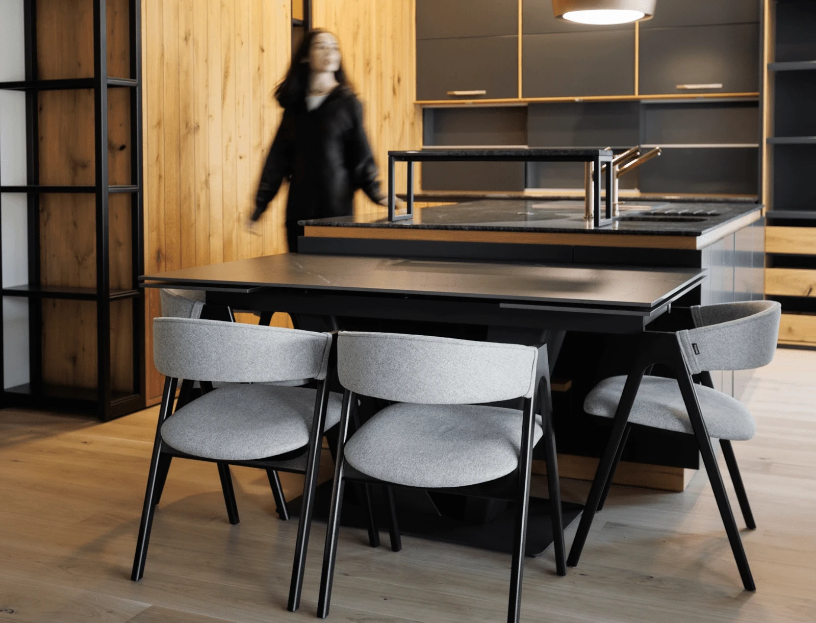 Four grey Lain chairs on black-stained ash frames around a dark dining table in a kitchen, woman walking past