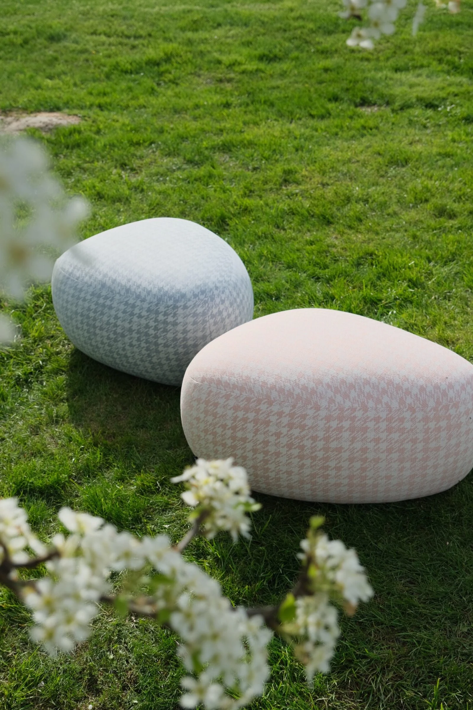 Two Chios poufs in pink and pale blue houndstooth fabric resting in a grassy garden with white blossoms