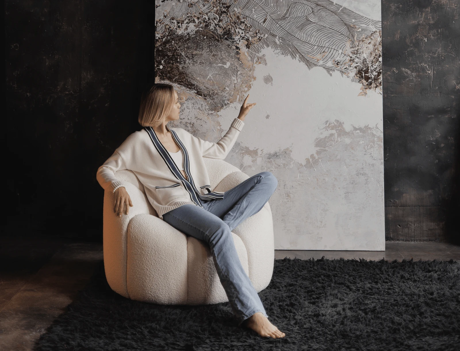 Model with a cream sweater seated on a cream Flora chair in a moody dark-walled room with an abstract framed artwork behind