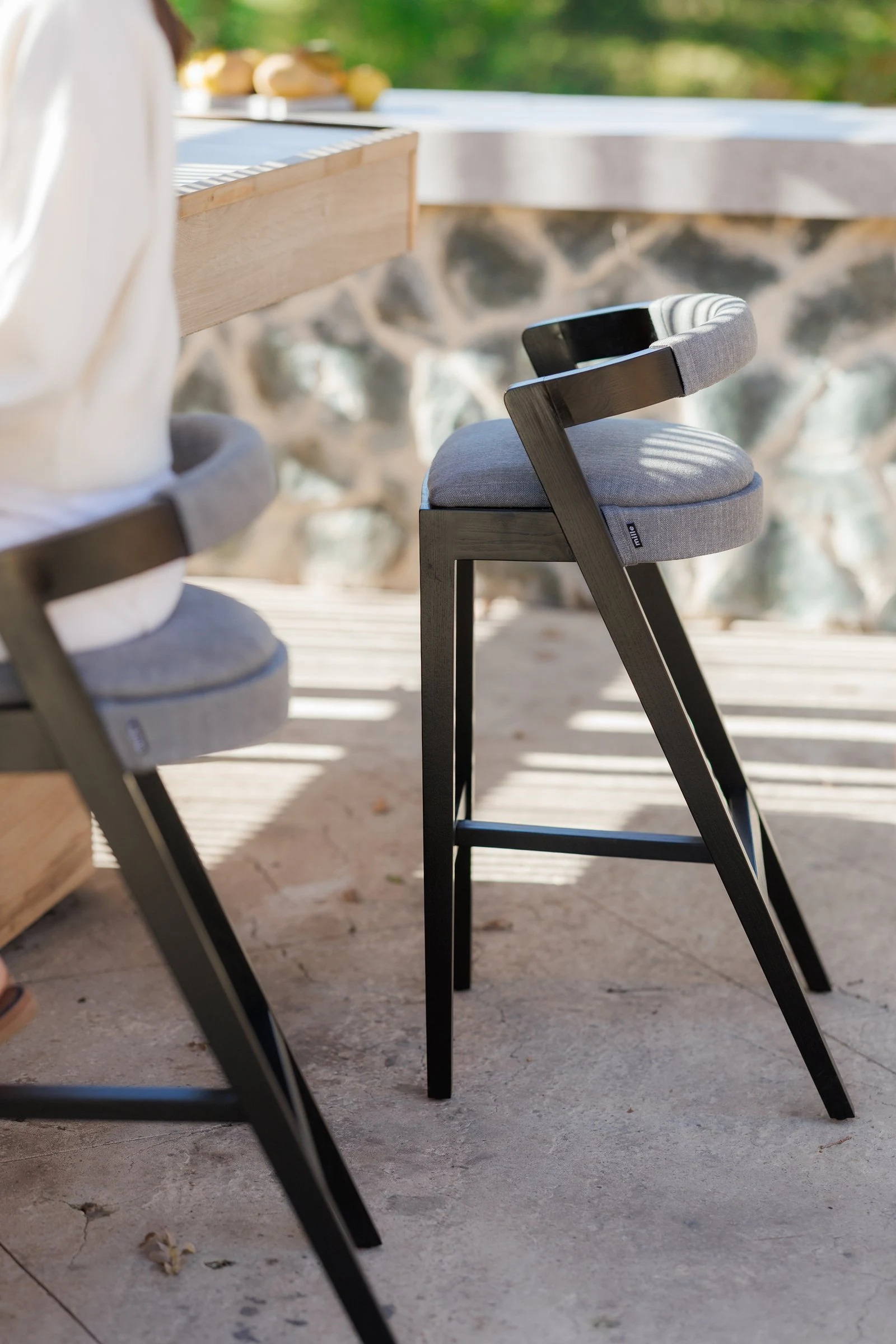 Side angle of a Baffi bar stool at the outdoor villa counter, black-stained ash frame and grey upholstered seat with stone wall behind