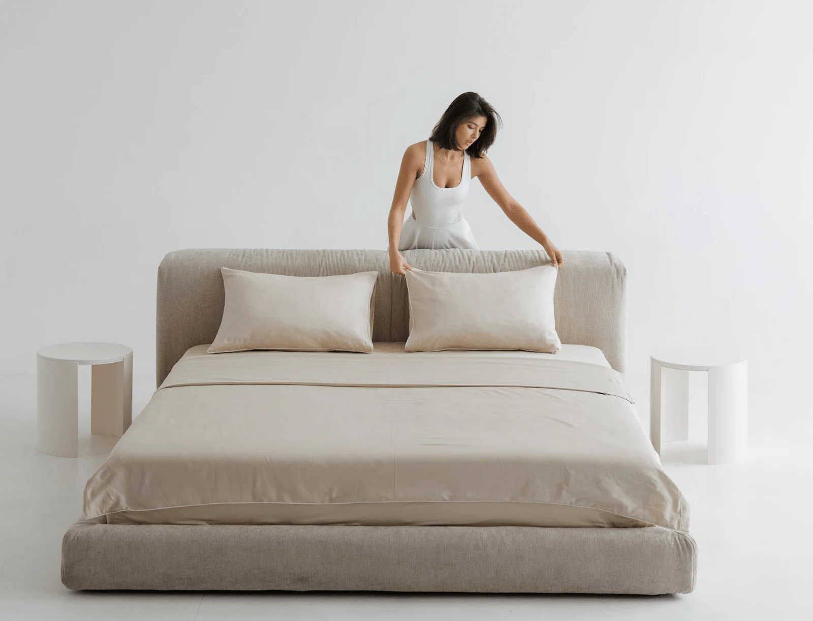 A woman in a white slip dress arranging pillows on a sand Grenoble 904 Marmaris bed in a bright minimal studio with two pebble side tables