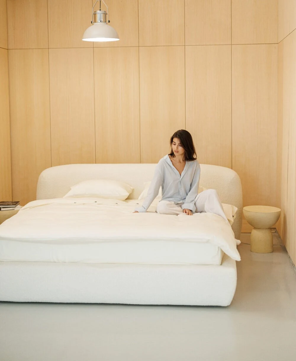 Cream boucle Belagio in a warm wood-paneled bedroom with model in pale blue pajamas sitting on the bed, books on a stone side table