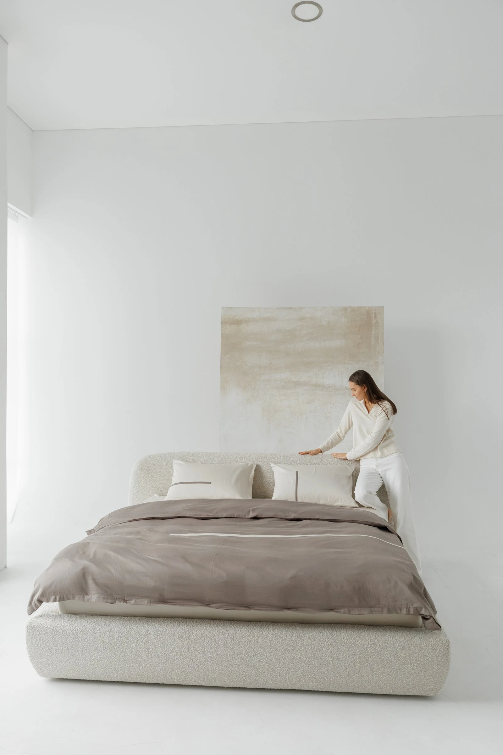 Cream boucle Belagio in a white studio with a painted canvas backdrop, model in cream tucking a brown linen sheet over the bed