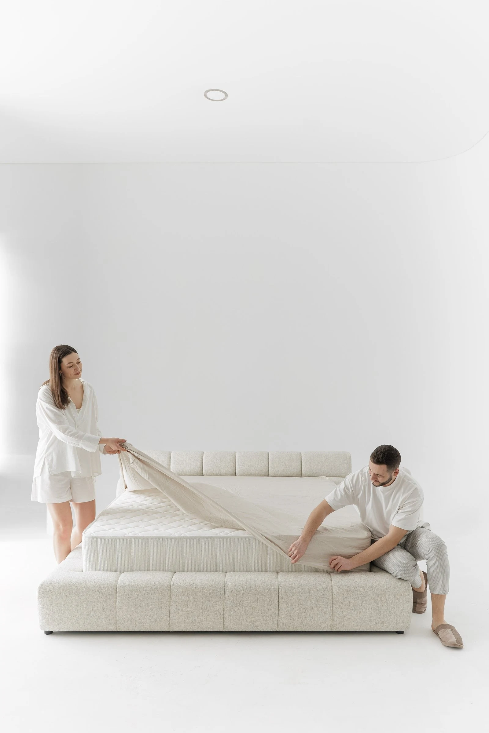 Grey tweed Nao mid-dressing, a couple fitting the sheet onto the bare mattress