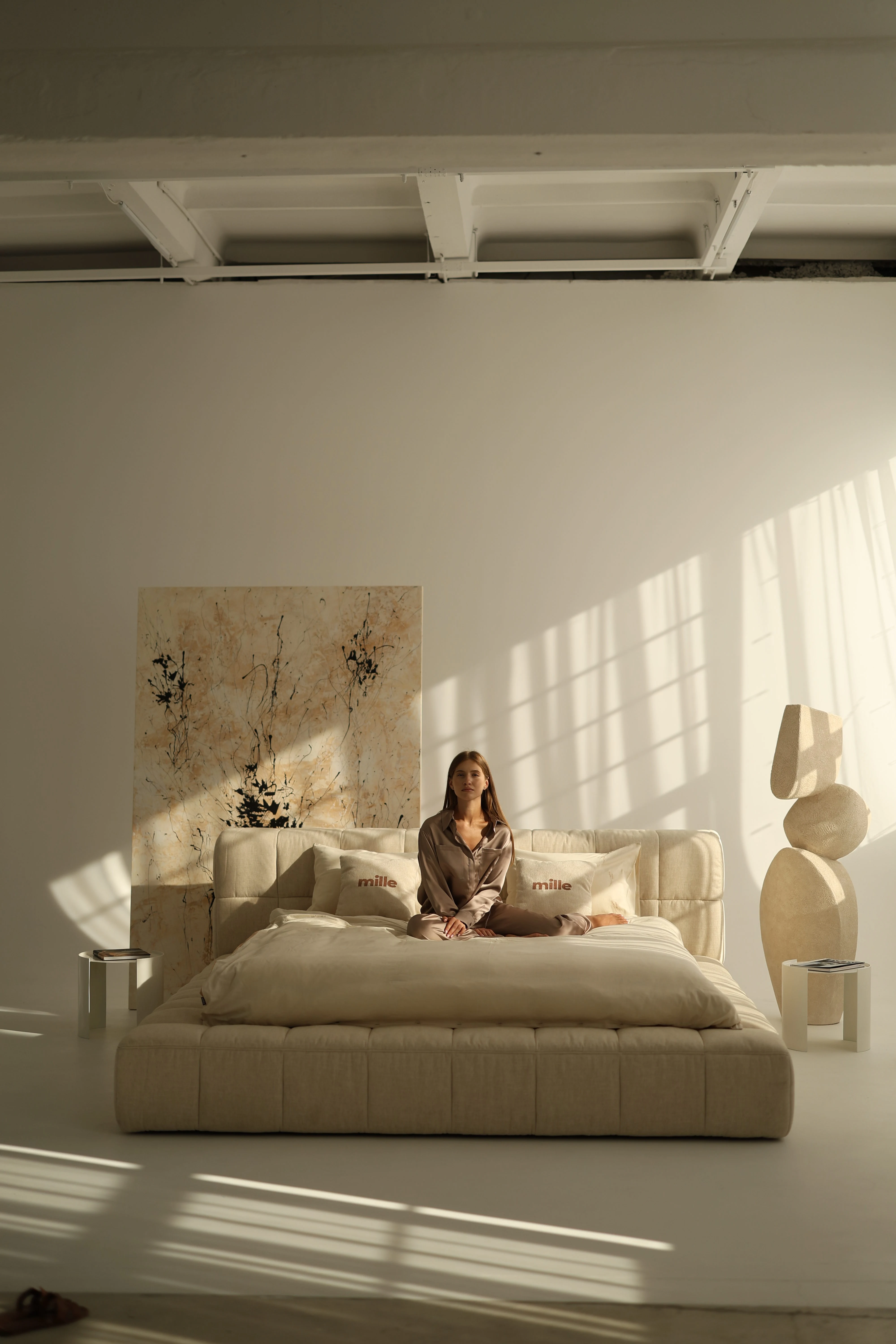 Sandy-fabric Nao bed in a warm loft with a model in silk loungewear seated on the made bed