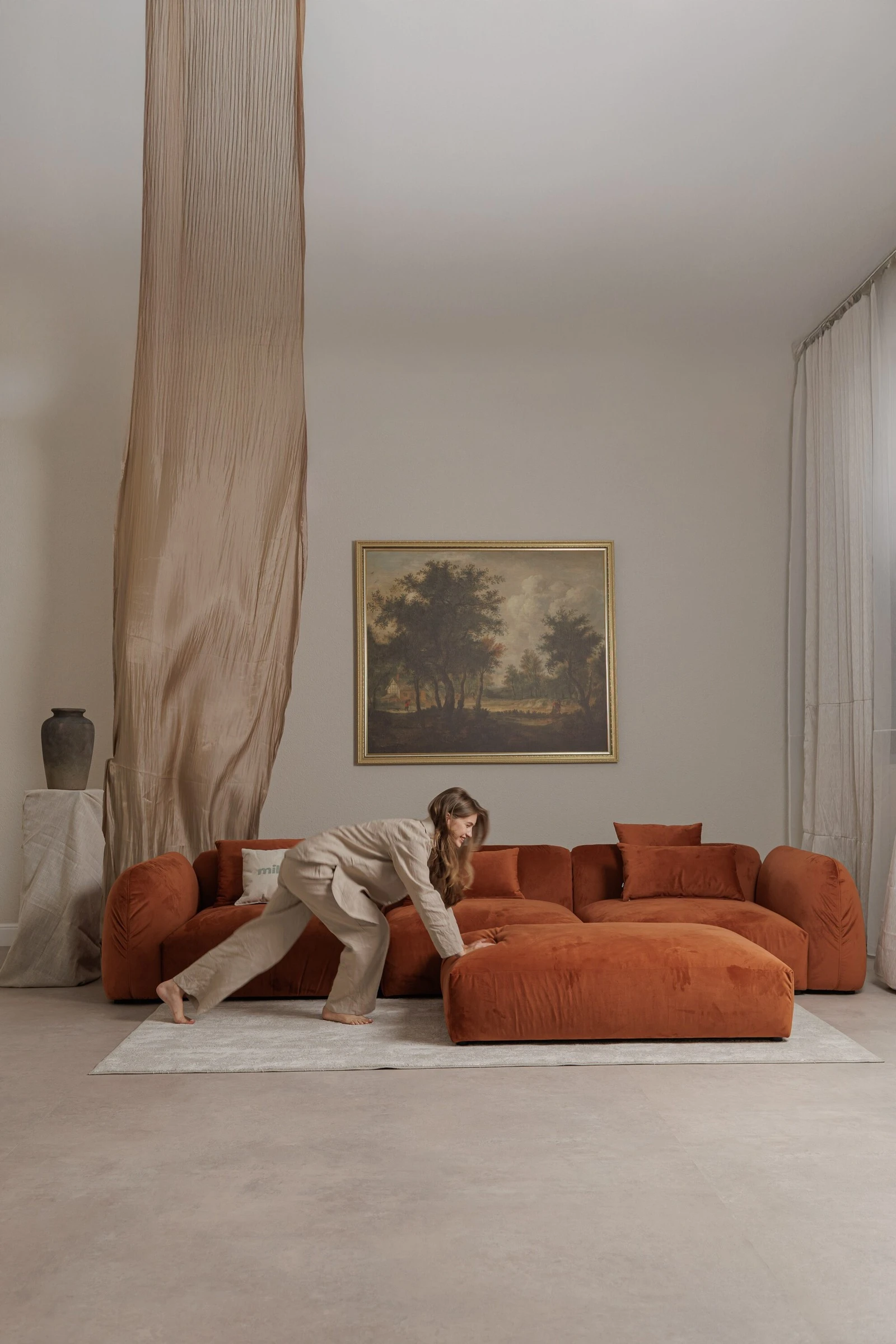 Rust velvet Tesa with model in linen reaching for the ottoman, gilt-framed landscape painting on the wall, sheer curtain behind