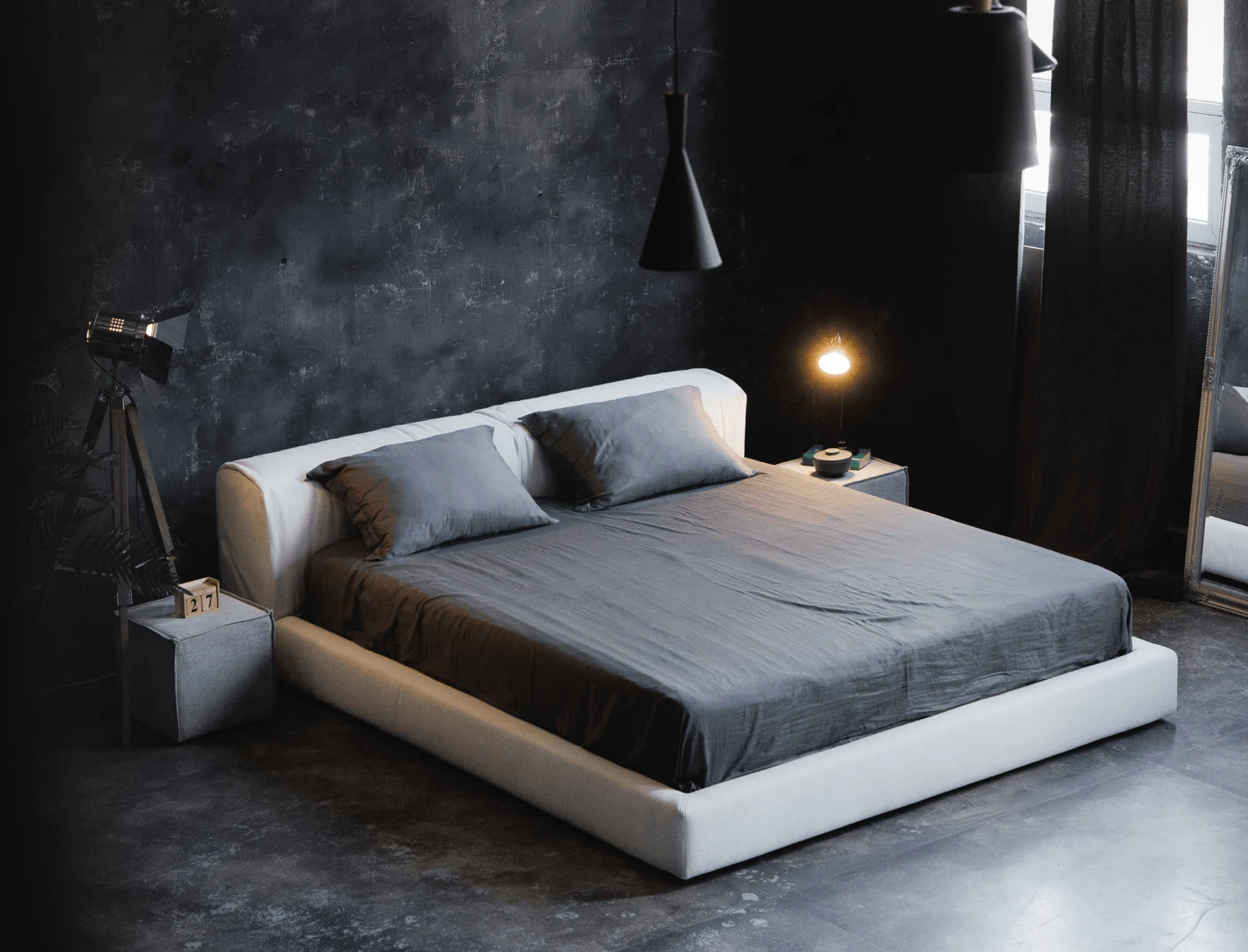Cream Soho 900 Marmaris bed in a dark concrete-walled loft with a brown linen blanket draped over the foot, a vintage camera tripod and pendant lamp in frame