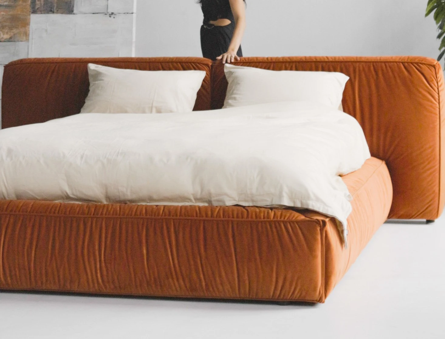 Burnt-orange Quartz 301 velvet Palermo bed in a loft with industrial windows, model in a black dress seated on the bed, white duvet draped across the velvet base