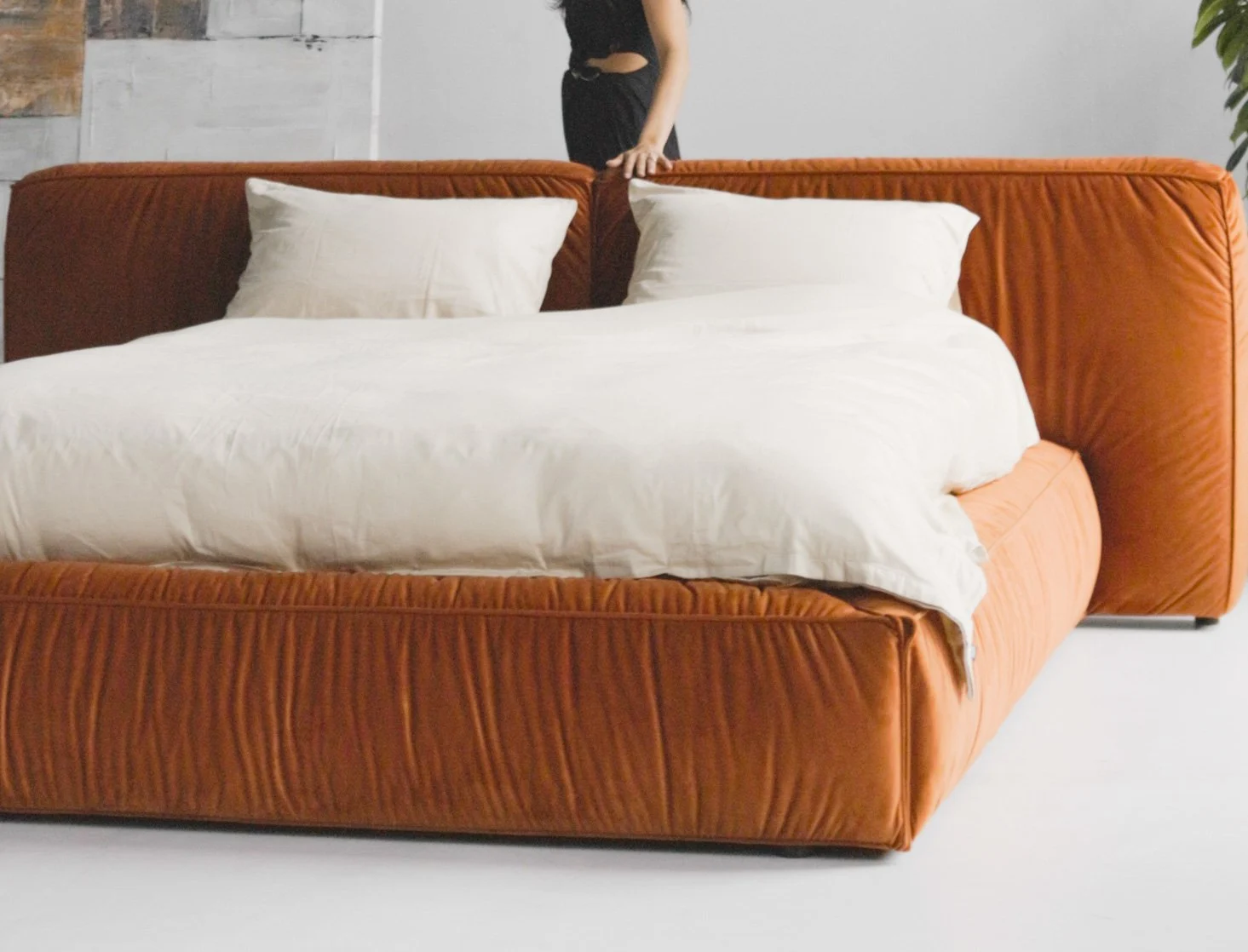 Burnt-orange Quartz 301 velvet Palermo bed in a loft with industrial windows, model in a black dress seated on the bed, white duvet draped across the velvet base