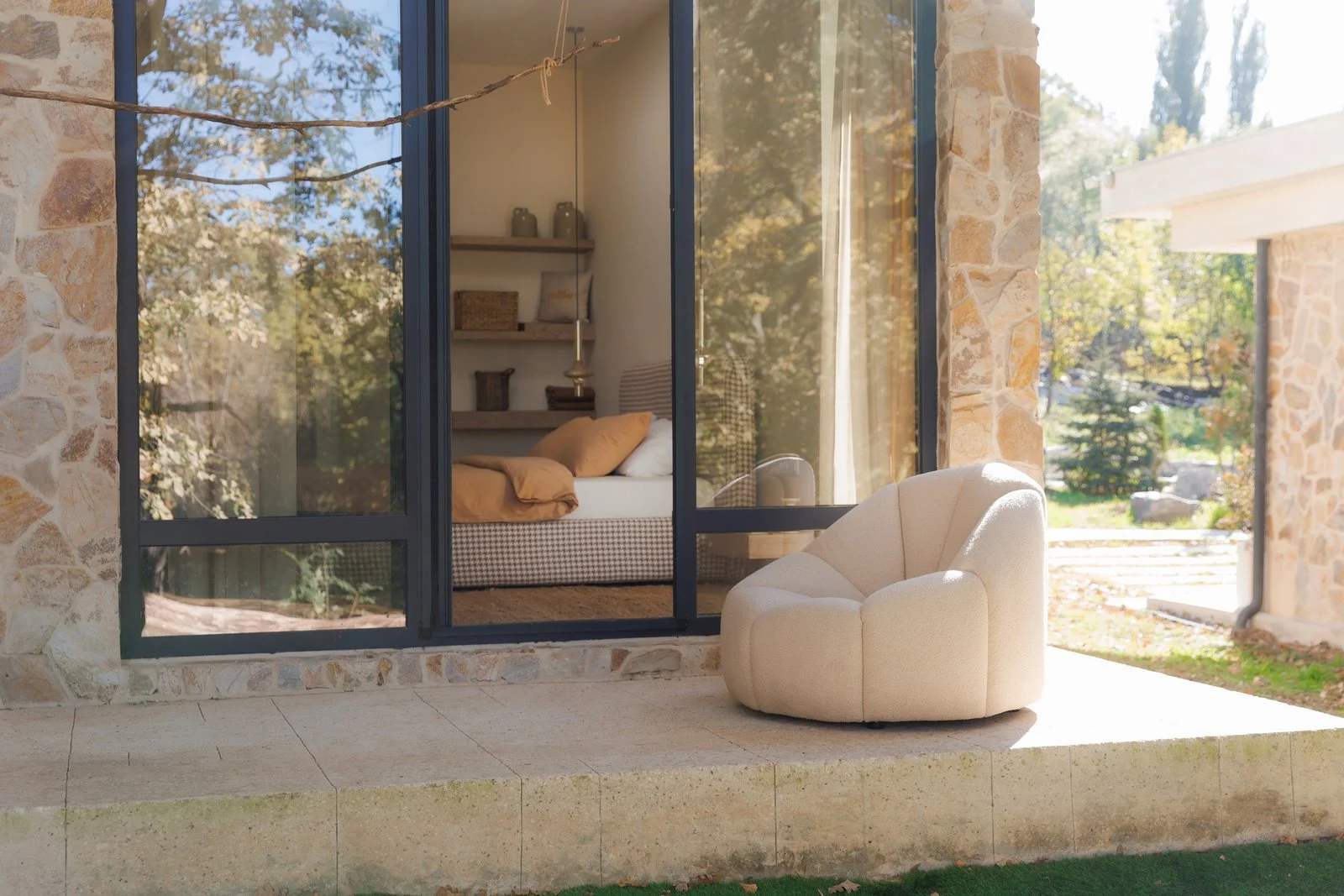 Cream boucle Flora lounge chair on a stone-walled villa patio with golden autumn trees and a bedroom visible through the glass behind