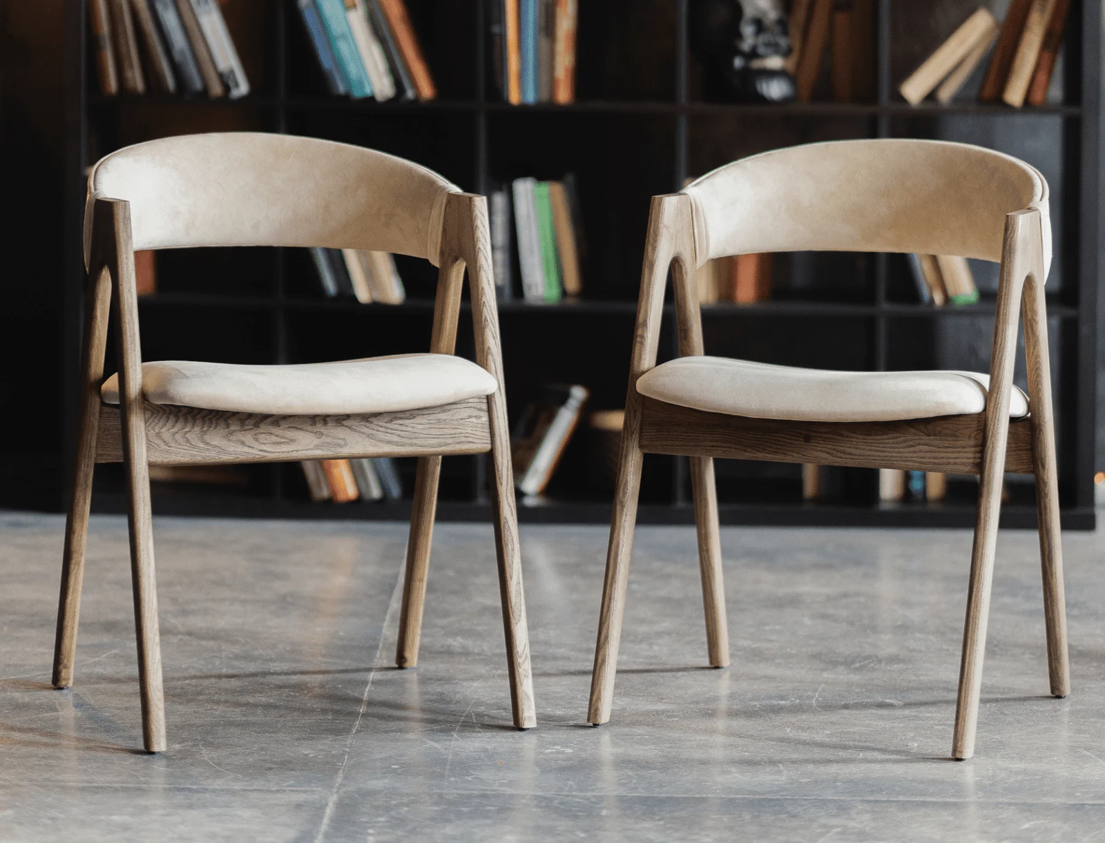 A pair of Lain dining chairs in a library room, light beige Bolzano fabric on a natural-ash sawhorse frame, against shelves filled with books