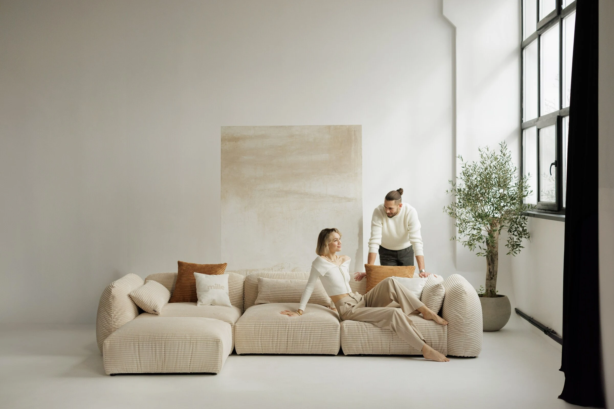 Tesa modular sofa with two figures arranging the cushions, daylight pouring in through a tall window