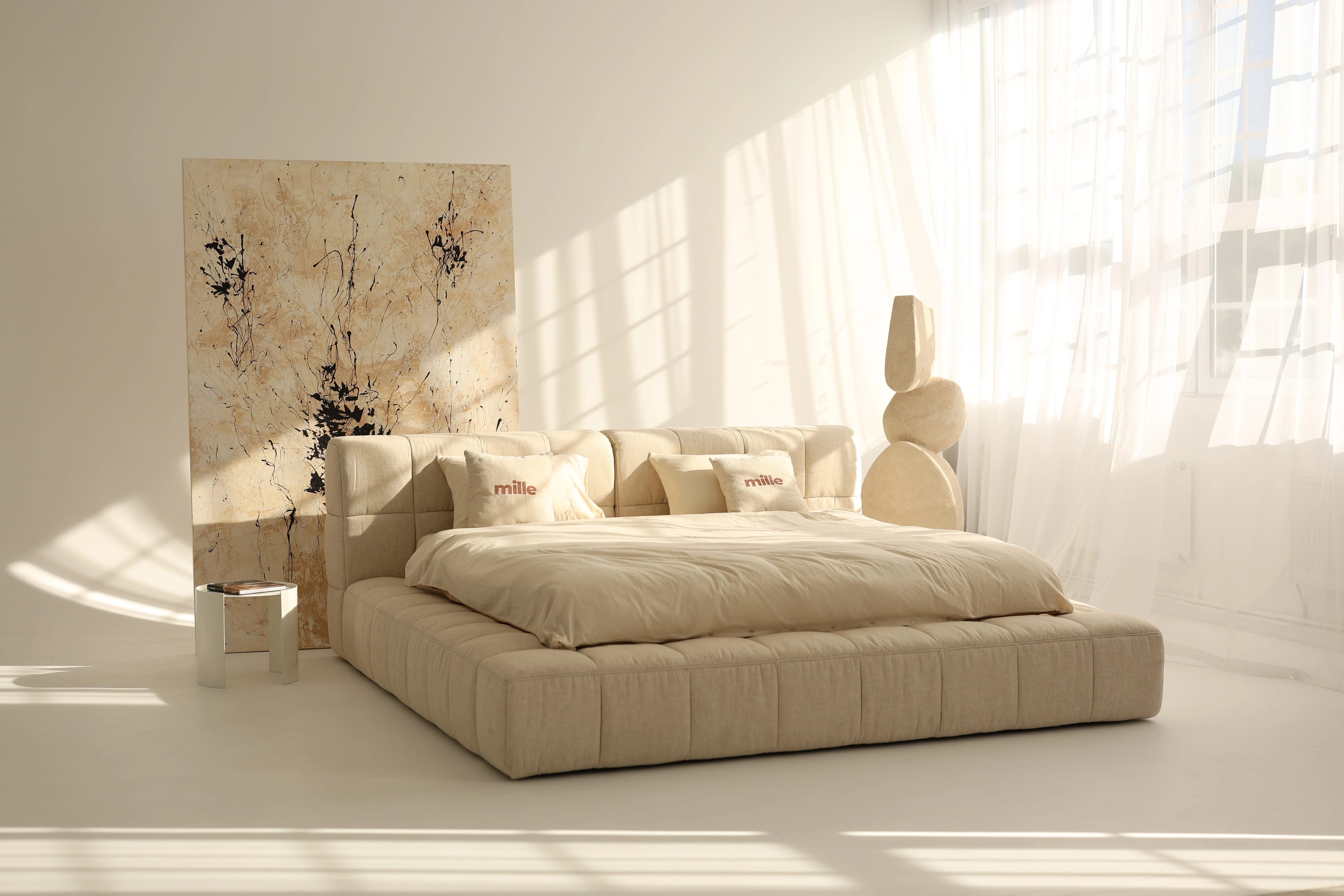 Sandy linen-channelled Nao bed in a warm sunlit loft with an abstract art panel and sculptural pieces nearby, model in silk loungewear seated on the bed