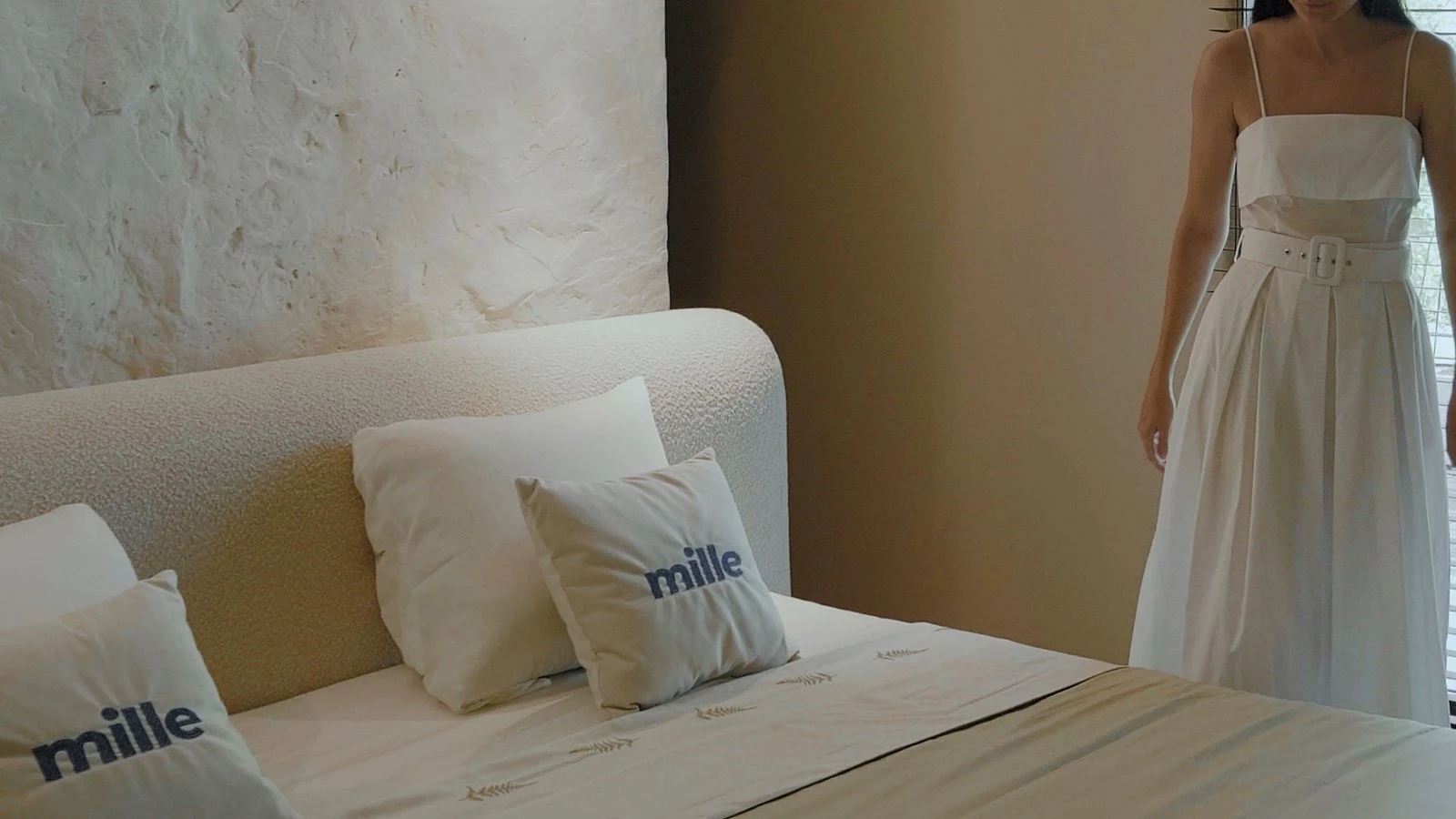 Editorial frame of the cream Fresco bed with a woman in a white dress arranging Mille-branded pillows on the bed