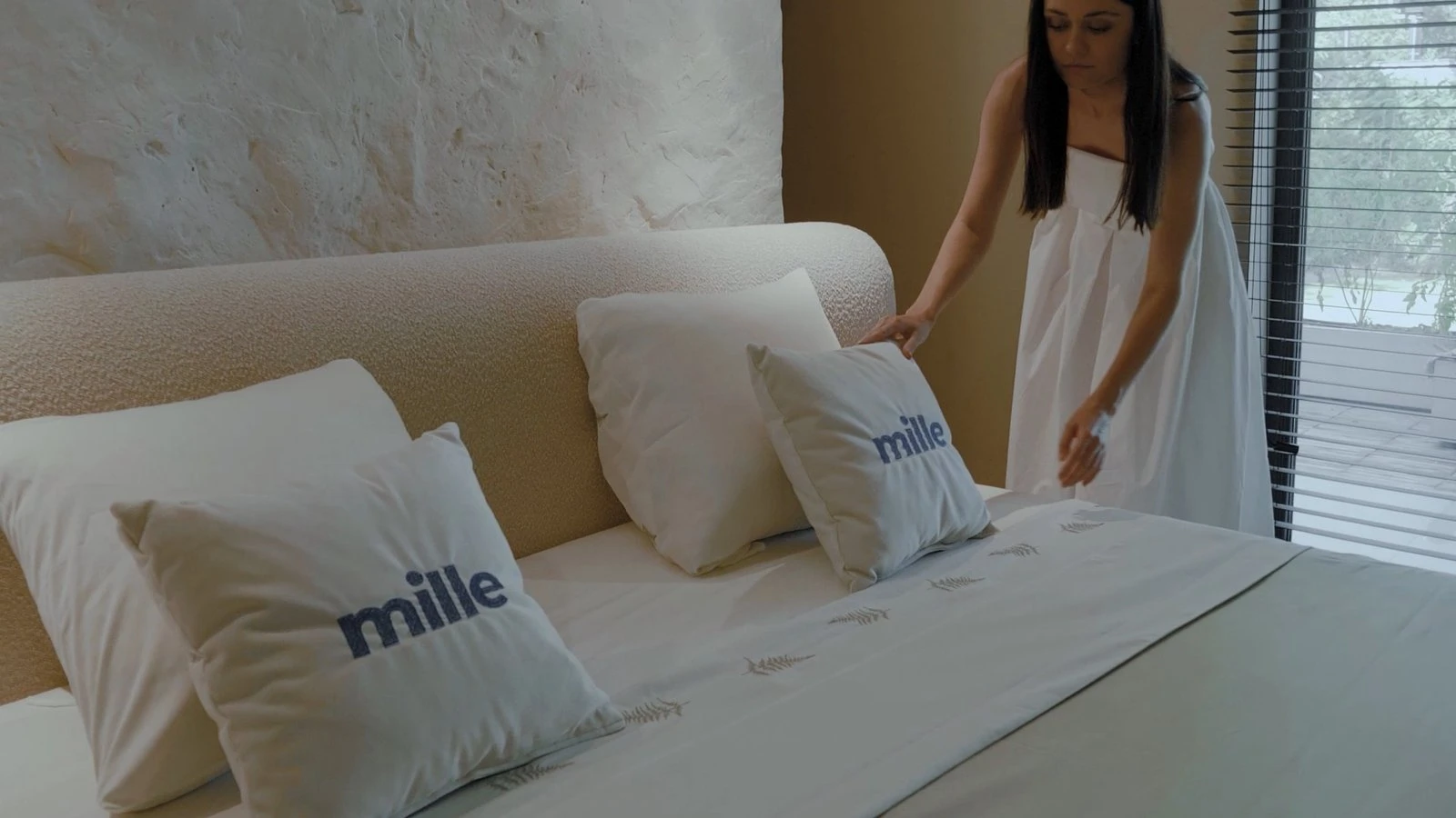 Woman in a white dress arranging Mille-branded pillows on the cream Fresco bed against a textured plaster wall
