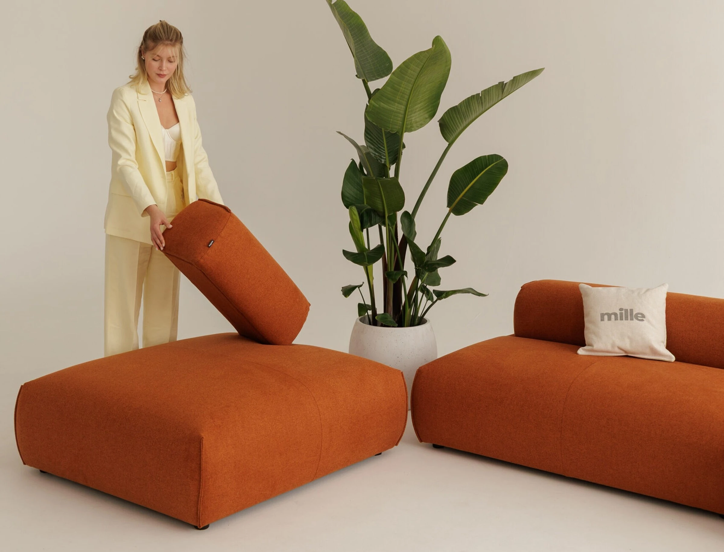 Model in cream on the rust Nissa holding the backrest cushion mid-rearrangement