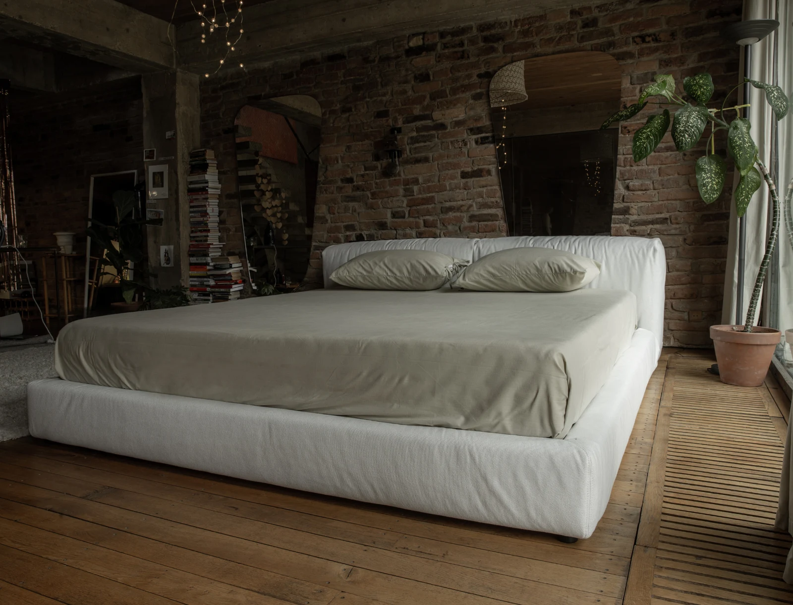 Cream Marmaris bed in a rustic brick loft with a vintage mirror, a plant, and a large window opening onto a sunny garden